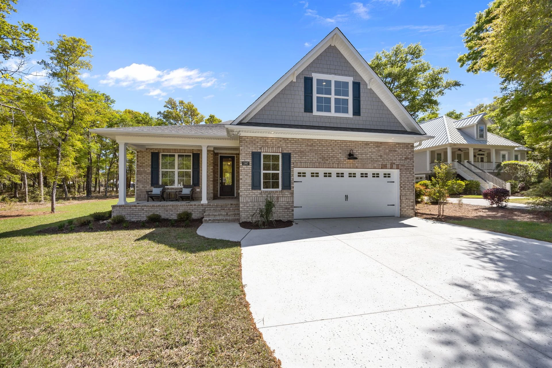 Modern brick home with a gabled roof, front porch, and two-car garage, set amidst green trees; ideal design for a home builder.
